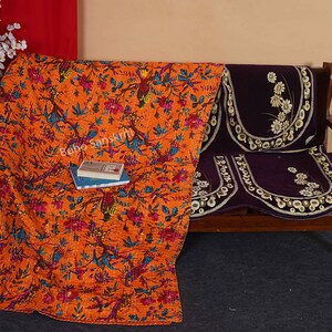 May include: Orange and blue floral kantha quilt with a book on top, draped over a brown wooden sofa with a purple floral cushion.