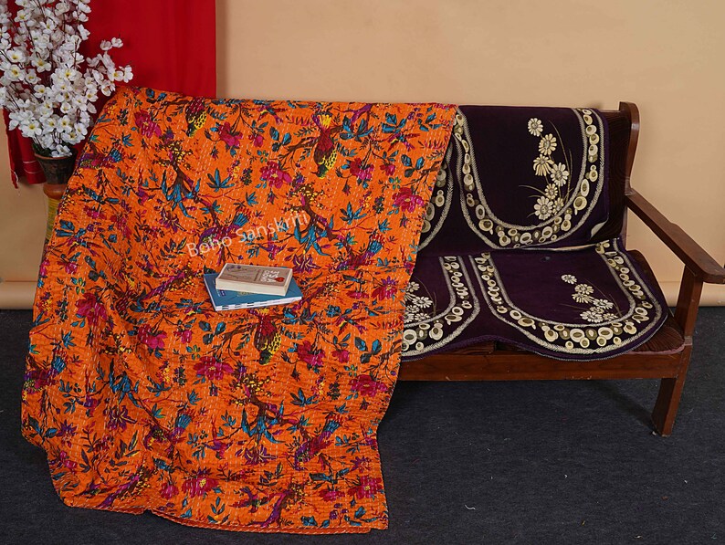 May include: Orange and blue floral kantha quilt with a book on top, draped over a brown wooden sofa with a purple floral cushion.