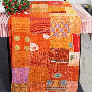 May include: A patchwork quilt featuring a mix of orange, red and purple fabrics. The quilt is made up of numerous fabric pieces sewn together to create a distinctive design. The quilt is folded and displayed on a table.