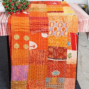Könnte beinhalten: Eine Patchwork-Decke mit verschiedenen Stoffen in Orange, Rot und Lila. Die Decke besteht aus vielen verschiedenen Stoffstücken, die zusammengenäht wurden, um ein einzigartiges Design zu schaffen. Die Decke ist gefaltet und auf einem Tisch ausgestellt.