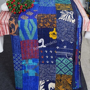 May include: A blue kantha quilt with a patchwork design featuring various colours and patterns. The quilt is made of recycled saris and is hand-stitched. The quilt is folded and displayed on a table.