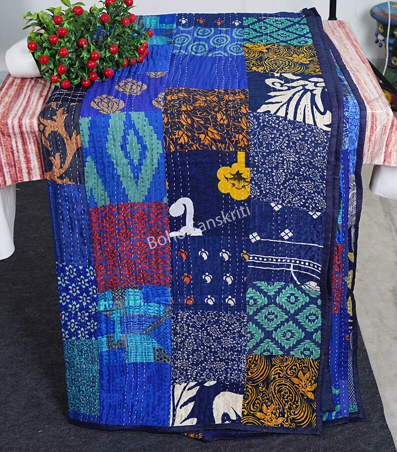 May include: A blue kantha quilt with a patchwork design featuring various colours and patterns. The quilt is made of recycled saris and is hand-stitched. The quilt is folded and displayed on a table.