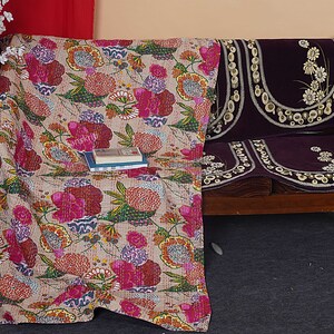 May include: A floral patterned kantha quilt in shades of pink, red, and green, draped over a wooden sofa with a purple velvet cushion cover. The cushion cover has a floral design with white embroidery.