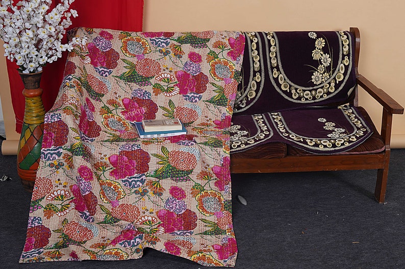 May include: A floral patterned kantha quilt in shades of pink, red, and green, draped over a wooden sofa with a purple velvet cushion cover. The cushion cover has a floral design with white embroidery.