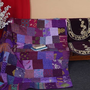 May include: A purple patchwork quilt with a variety of patterns and textures. The quilt is draped over a wooden couch with a purple floral patterned cushion.