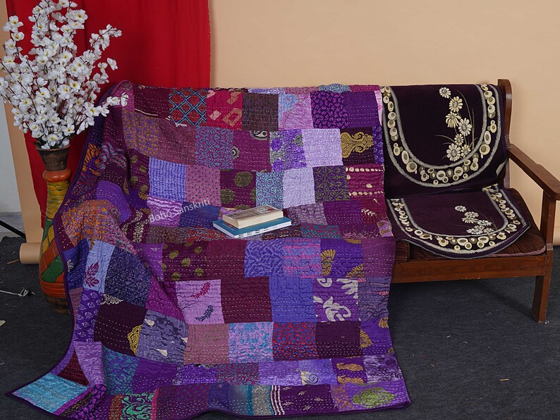 May include: A purple patchwork quilt with a variety of patterns and textures. The quilt is draped over a wooden couch with a purple floral patterned cushion.