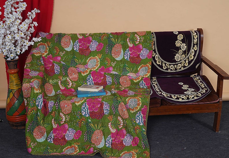 May include: A green kantha quilt with a floral and pineapple print. The quilt is draped over a wooden chair with a purple cushion.