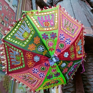 Puede incluir: Un paraguas colorido de varios paneles con un dise&ntilde;o verde, rosa y rojo. El paraguas est&aacute; decorado con flores bordadas, huellas de manos y elefantes. El paraguas tiene un ribete de flecos y una borla.