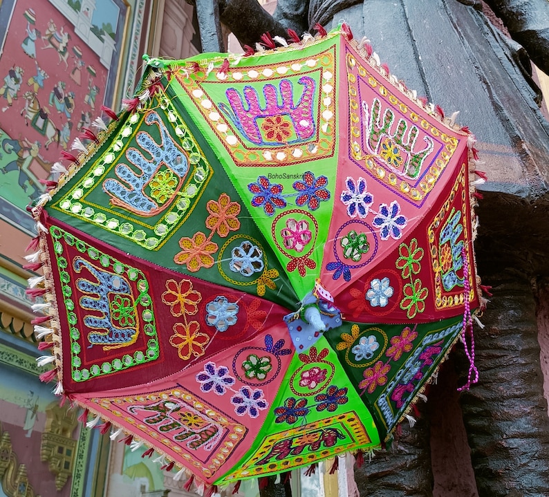 Puede incluir: Un paraguas colorido de varios paneles con un dise&ntilde;o verde, rosa y rojo. El paraguas est&aacute; decorado con flores bordadas, huellas de manos y elefantes. El paraguas tiene un ribete de flecos y una borla.