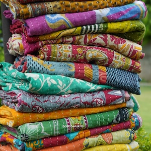 Könnte beinhalten: Ein Stapel bunter Kantha-Steppdecken mit komplizierten Blumenmustern. Die Steppdecken sind aus recycelten Saris hergestellt und handgenäht. Die Steppdecken sind in einem Stapel angeordnet, wobei die oberste Steppdecke umgeklappt ist. Die Steppdecken sind ein lebendiges Farbenspiel, darunter Rot, Orange, Gelb, Grün, Blau und Lila. Die Steppdecken sind ein wunderschönes Beispiel für traditionelles indisches Handwerk.