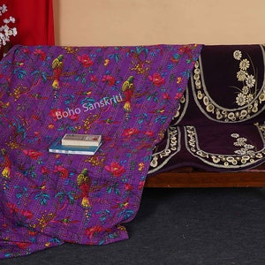 May include: Purple kantha quilt with a floral and bird pattern. The quilt is draped over a wooden couch with purple cushions. The text "boho sanskriti" is visible on the quilt.