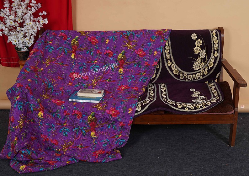 May include: Purple kantha quilt with a floral and bird pattern. The quilt is draped over a wooden couch with purple cushions. The text "boho sanskriti" is visible on the quilt.