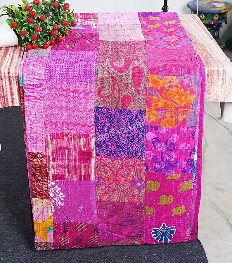 May include: A pink patchwork table runner made from silk fabric with various patterns. The runner is approximately 46 cm wide and 183 cm long.