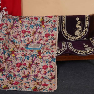 May include: A beige kantha quilt with a floral and bird pattern. The quilt is draped over a wooden sofa with a purple velvet cushion cover with embroidered white flowers.