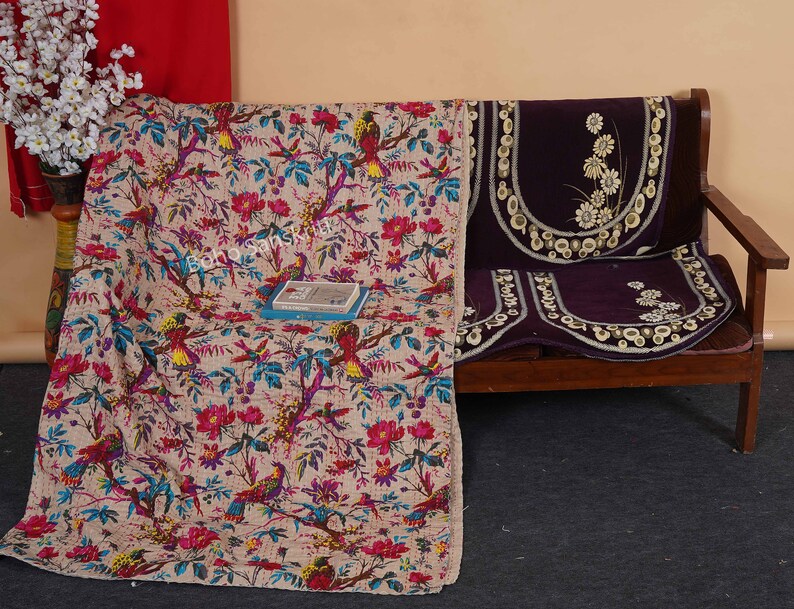 May include: A beige kantha quilt with a floral and bird pattern. The quilt is draped over a wooden sofa with a purple velvet cushion cover with embroidered white flowers.