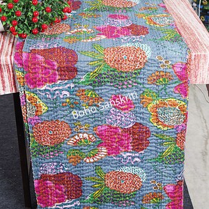 May include: A grey table runner with a floral pattern in shades of pink, orange, yellow, and green. The runner is quilted and has a "boho sanskrit" logo in the centre.