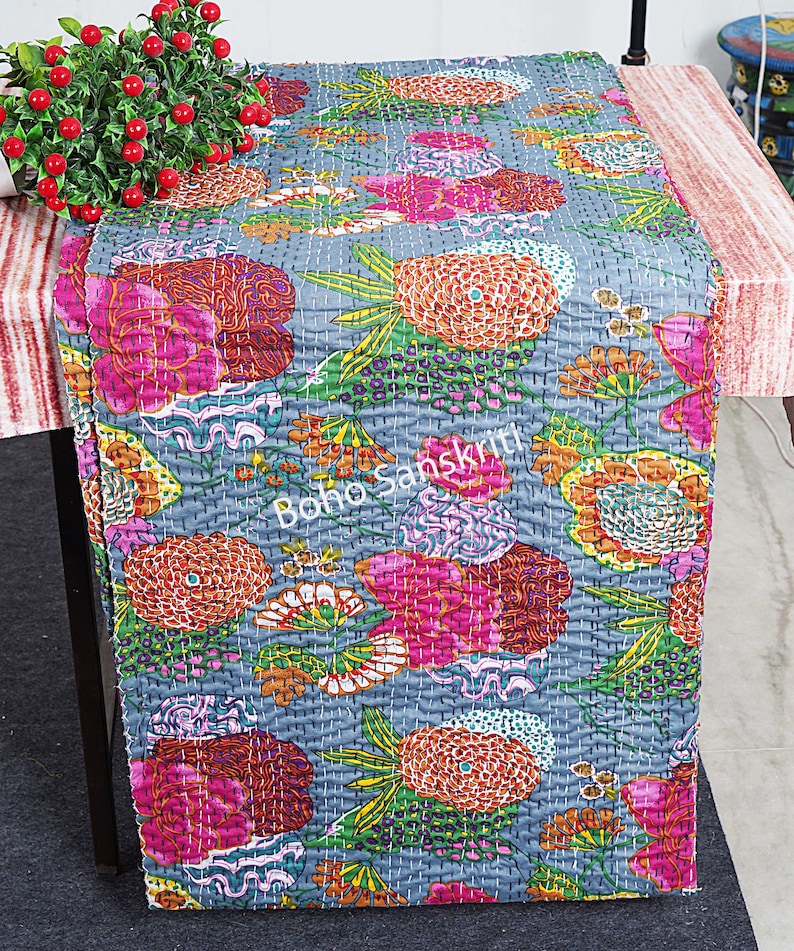 May include: A grey table runner with a floral pattern in shades of pink, orange, yellow, and green. The runner is quilted and has a "boho sanskrit" logo in the centre.