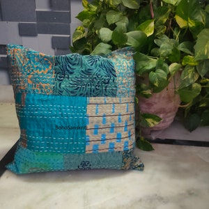 May include: Square patchwork pillow in shades of teal, blue, and beige with white stitching. The pillow features a variety of patterned fabric pieces, including floral and geometric designs. The text "BohoSanskriti" is visible on the pillow.