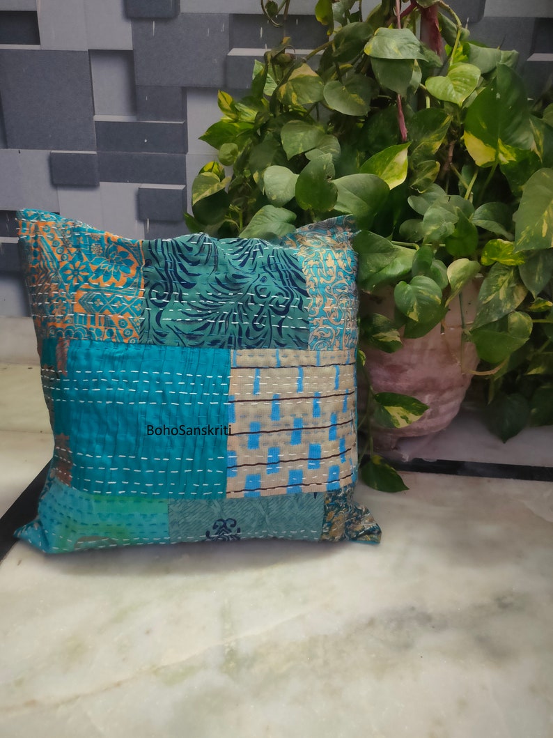 May include: Square patchwork pillow in shades of teal, blue, and beige with white stitching. The pillow features a variety of patterned fabric pieces, including floral and geometric designs. The text "BohoSanskriti" is visible on the pillow.