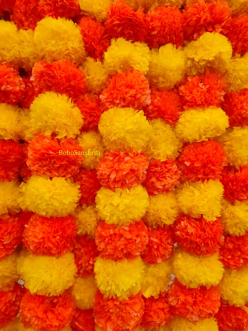 May include: A garland of yellow and orange paper marigolds, also known as marigold pom poms, arranged in a repeating pattern. The garland is a traditional Indian decoration used for festivals and celebrations.