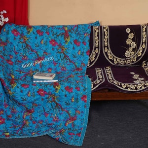 May include: A turquoise blue kantha quilt with a floral and bird print. The quilt is folded and draped over a wooden bench with a dark purple cushion. The text "boho sanskriti" is visible on the quilt.