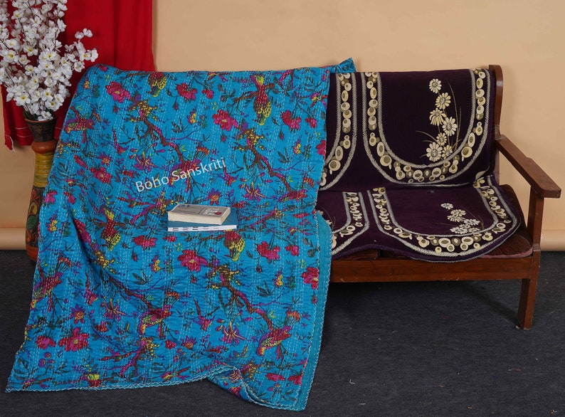 May include: A turquoise blue kantha quilt with a floral and bird print. The quilt is folded and draped over a wooden bench with a dark purple cushion. The text "boho sanskriti" is visible on the quilt.