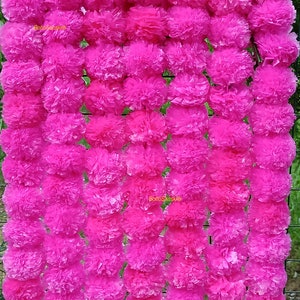 May include: A garland of pink paper marigolds, also known as marigold flowers, strung together. The garland is made of many individual marigolds, each with a fluffy, layered texture. The garland is hanging on a wire fence.