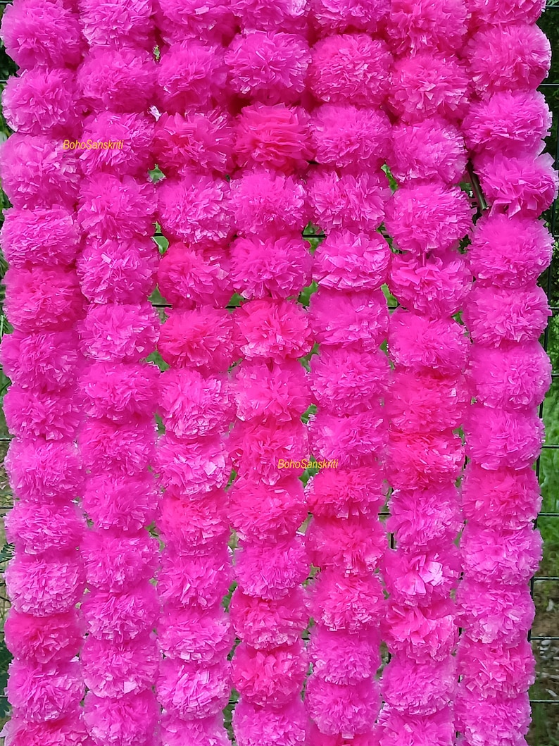 May include: A garland of pink paper marigolds, also known as marigold flowers, strung together. The garland is made of many individual marigolds, each with a fluffy, layered texture. The garland is hanging on a wire fence.