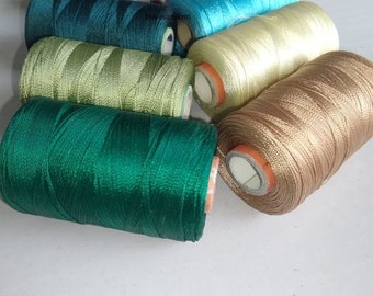 May include: A collection of spools of thread in various colors, including green, yellow, blue, red, and brown. The spools are arranged in a semi-circle on a white surface.