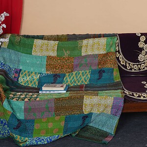 May include: A colorful patchwork quilt made of recycled saris, draped over a wooden couch with a floral patterned cushion. The quilt has a variety of colors, including green, blue, purple, and brown.