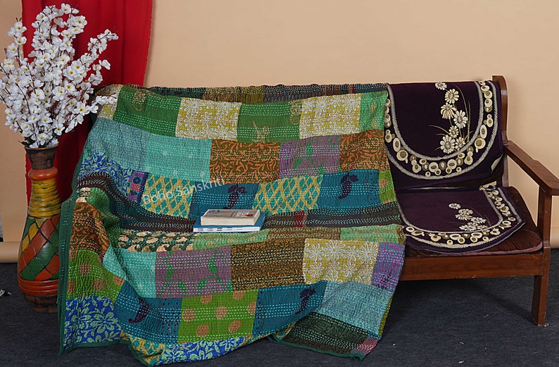 May include: A colorful patchwork quilt made of recycled saris, draped over a wooden couch with a floral patterned cushion. The quilt has a variety of colors, including green, blue, purple, and brown.