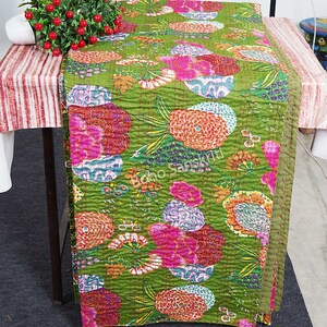May include: A green kantha quilt with a floral pattern in shades of pink, orange, and purple. The quilt is folded and displayed on a table.