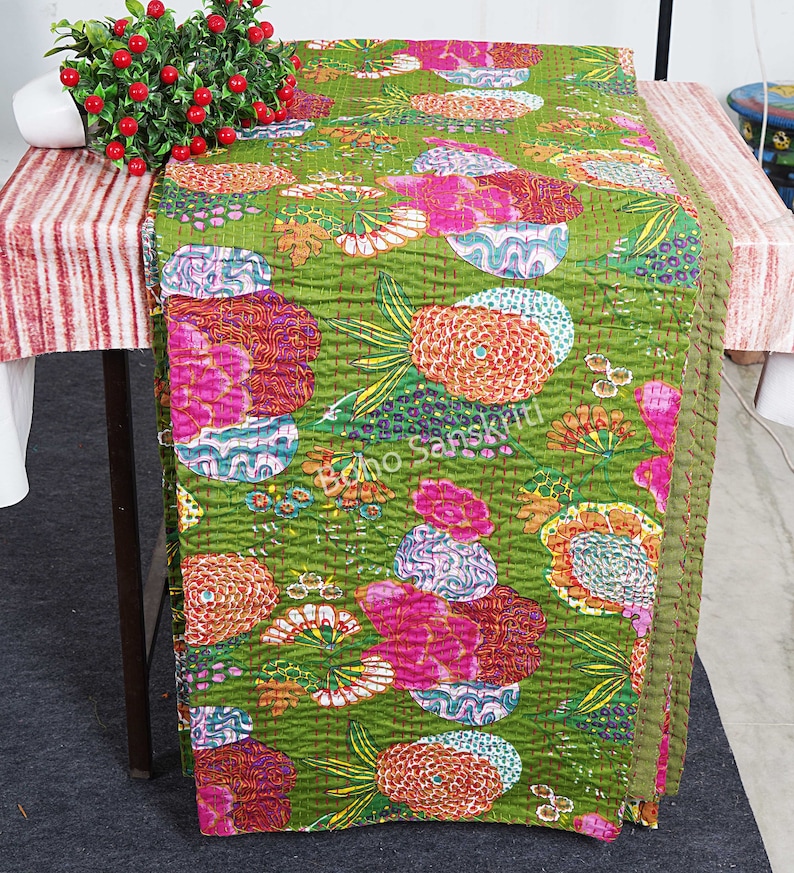May include: A green kantha quilt with a floral pattern in shades of pink, orange, and purple. The quilt is folded and displayed on a table.