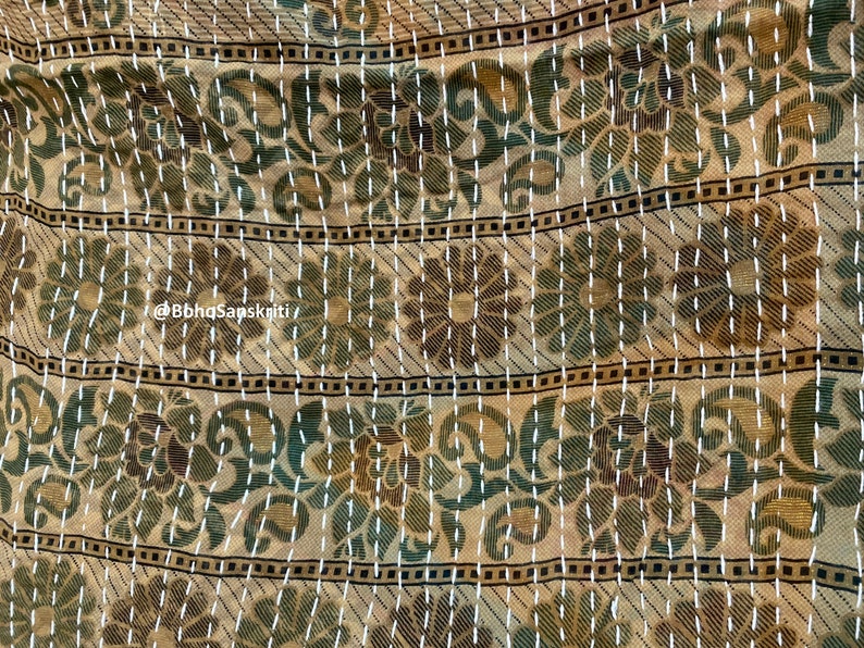 May include: A beige fabric with a repeating pattern of green and brown flowers. The fabric is stitched with white thread in a running stitch pattern. The fabric has a subtle sheen and a soft texture.