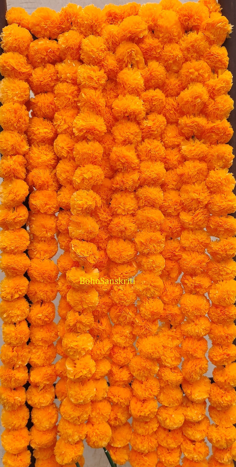 May include: A garland of bright orange marigold flowers, strung together in a long strand. The flowers are made of paper and are arranged in a repeating pattern.