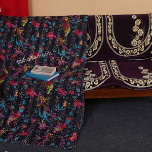 May include: A black kantha quilt with a floral and bird pattern, draped over a wooden bench with a purple velvet cushion cover. The cushion cover has a floral design with white embroidery.