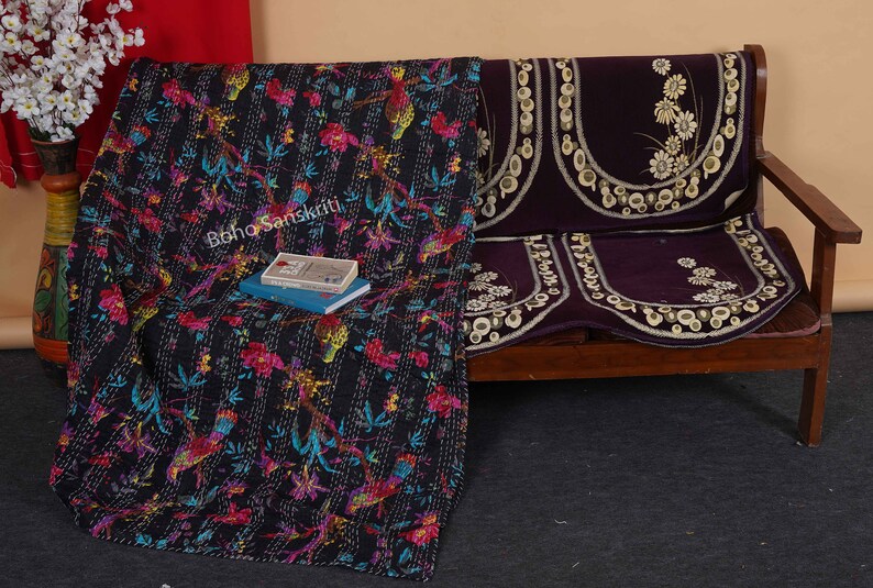 May include: A black kantha quilt with a floral and bird pattern, draped over a wooden bench with a purple velvet cushion cover. The cushion cover has a floral design with white embroidery.