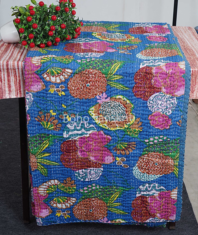 May include: A blue table runner with a floral pattern in shades of pink, orange, green, and brown. The runner is made of cotton and has a quilted texture.