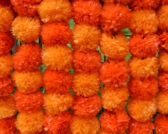 Op de afbeelding: Een close-up van een muur van levendige oranje goudsbloembloemen. De bloemen zijn dicht opeengepakt, waardoor een textuur ontstaat. Het kleurverloop varieert van lichtoranje tot dieporanje, met een paar groene bladeren zichtbaar.