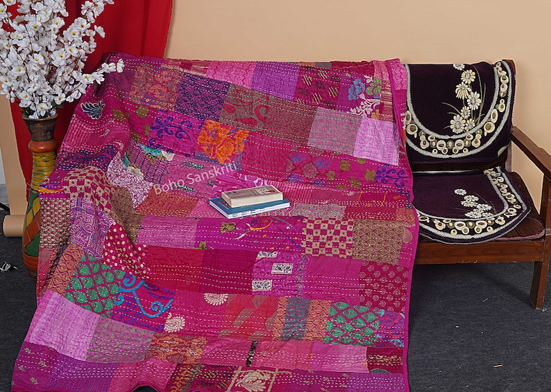 May include: A pink kantha quilt with a patchwork design. The quilt is made of various fabrics and is hand-stitched. The quilt is folded and placed on a wooden chair with a purple cushion. The text "boho sanskriti" is visible on the quilt.