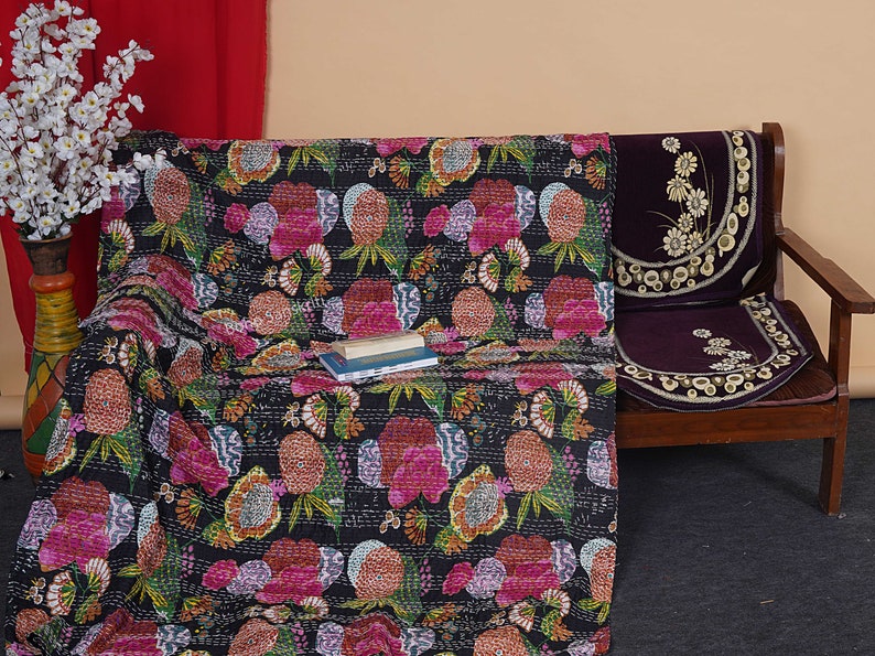 May include: A black kantha quilt with a floral pattern in shades of pink, orange, and green. The quilt is draped over a wooden chair with a purple cushion.