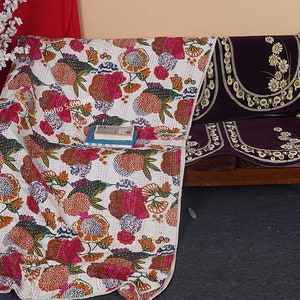 May include: A white kantha quilt with a floral pattern in shades of pink, orange, and green. The quilt is draped over a wooden bench with purple cushions featuring embroidered floral designs.