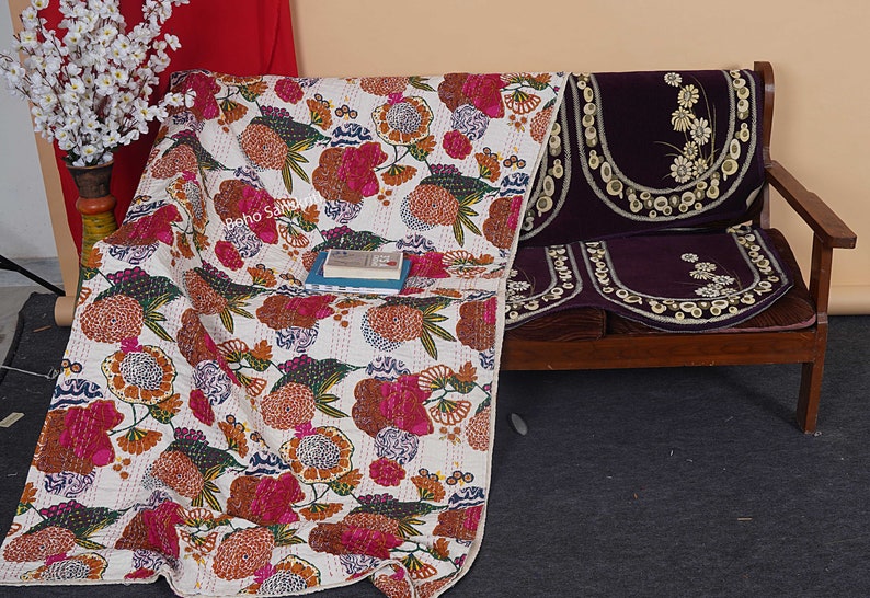 May include: A white kantha quilt with a floral pattern in shades of pink, orange, and green. The quilt is draped over a wooden bench with purple cushions featuring embroidered floral designs.