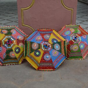 May include: Three colorful, decorative umbrellas with intricate patterns and embellishments. Each umbrella features a unique design with vibrant hues of red, yellow, blue, and green, adorned with reflective accents and a fringed edge. The umbrellas are displayed on a stone surface.