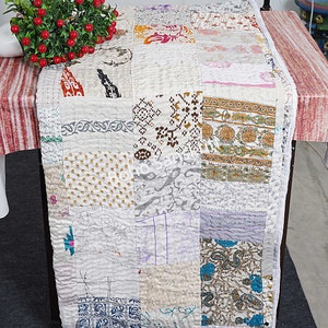 May include: A patchwork table runner with a variety of patterned fabric squares in cream, white, and pastel colors. The runner features floral, paisley, and geometric designs. The edges are finished with a white border, creating a textured, handcrafted look.