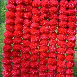 May include: A garland of bright red marigold flowers, made of fabric, arranged in a vertical row. The garland is hanging on a wire fence. The text "Boho Sanskriti" is visible on the garland.
