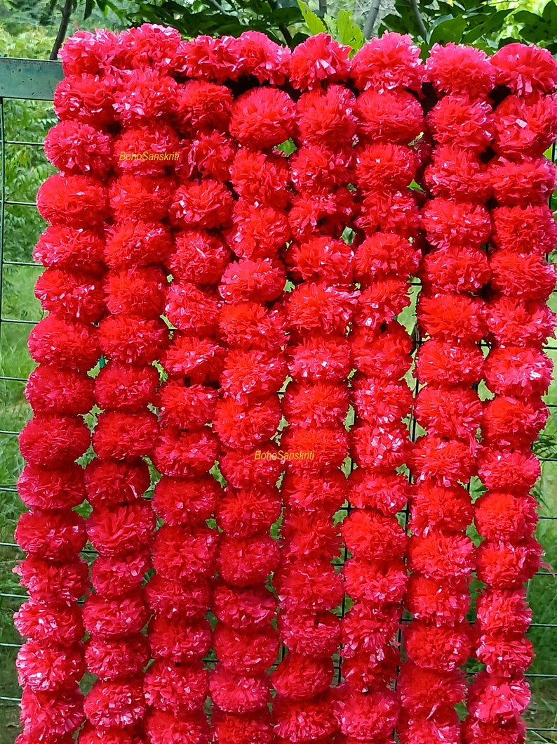 May include: A garland of bright red marigold flowers, made of fabric, arranged in a vertical row. The garland is hanging on a wire fence. The text "Boho Sanskriti" is visible on the garland.