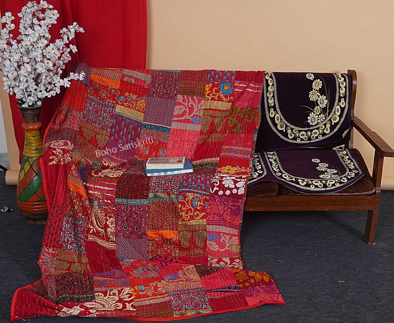 May include: A red, orange, and purple patchwork quilt with a floral pattern. The quilt is folded and draped over a wooden couch with a purple cushion. The text "Boho Sanskriti" is visible on the quilt.
