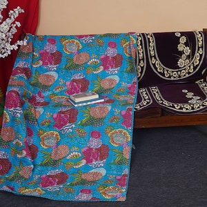 May include: A turquoise blue kantha quilt with a floral pattern in shades of pink, orange, yellow, and green. The quilt is draped over a wooden bench with a purple velvet cushion.