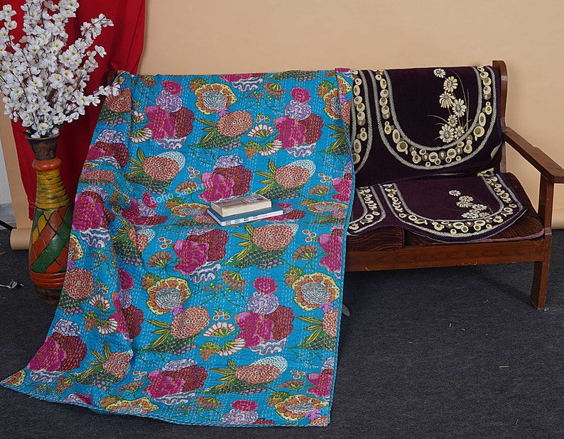 May include: A turquoise blue kantha quilt with a floral pattern in shades of pink, orange, yellow, and green. The quilt is draped over a wooden bench with a purple velvet cushion.